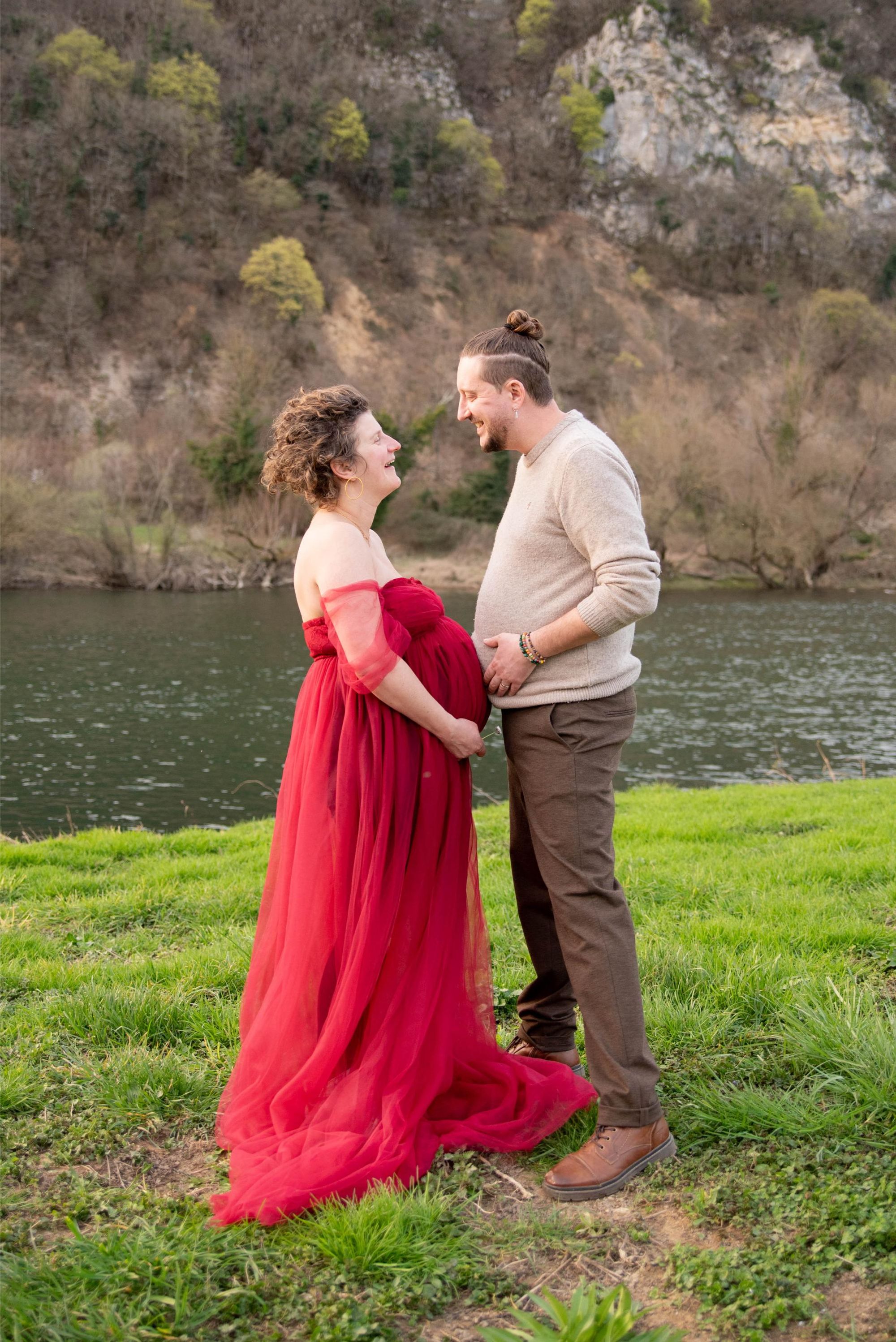 photo de 2 futurs parents ventre rond, lors d'une séance photo grossesse. Le papa plaisante en imitant à future maman, séance en extérieur au bord du doubs à Besançon