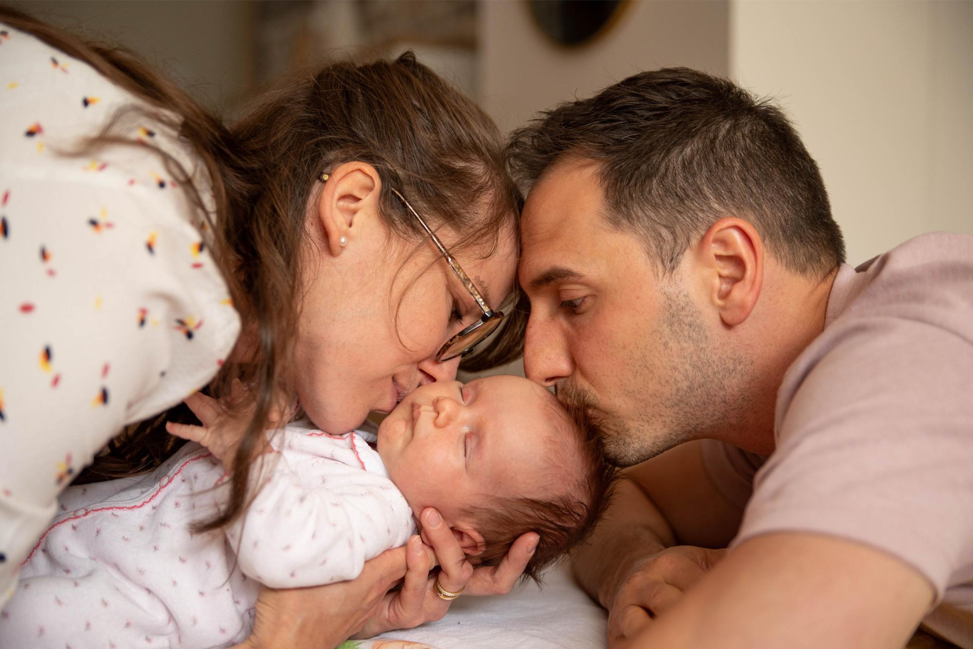 photo de 2 jeunes parents embrassant leur bébé lors d'une session naissance intimiste réalisée à domicile proche de Besançon